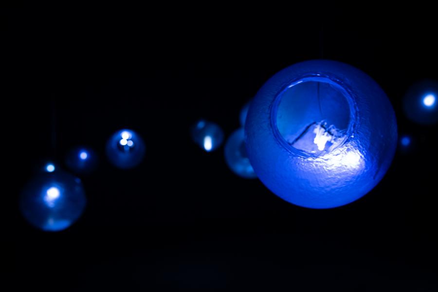A dark gallery space with numerous glowing blue glass orbs suspended from the ceiling, reflecting their light on the polished floor below.