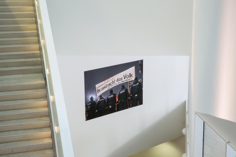 A photograph installed on the wall of a stairwell, viewed from an elevated perspective. The image features police officers in riot gear, facing away, with a banner in German that translates to "You are not the people." The white walls and minimal architecture frame the piece.
