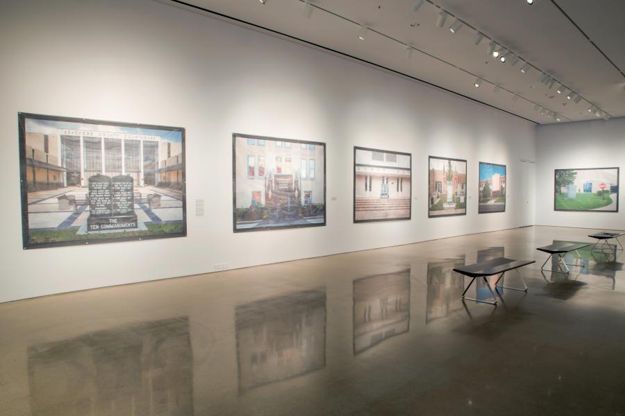 Gallery installation view featuring large-scale photographs of various courthouse buildings displayed on white walls, with polished concrete floors reflecting the artworks and benches arranged for viewers.