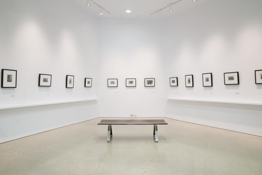 A minimalist gallery space featuring framed black-and-white photographs evenly spaced on white walls, with a simple bench centred in the room.