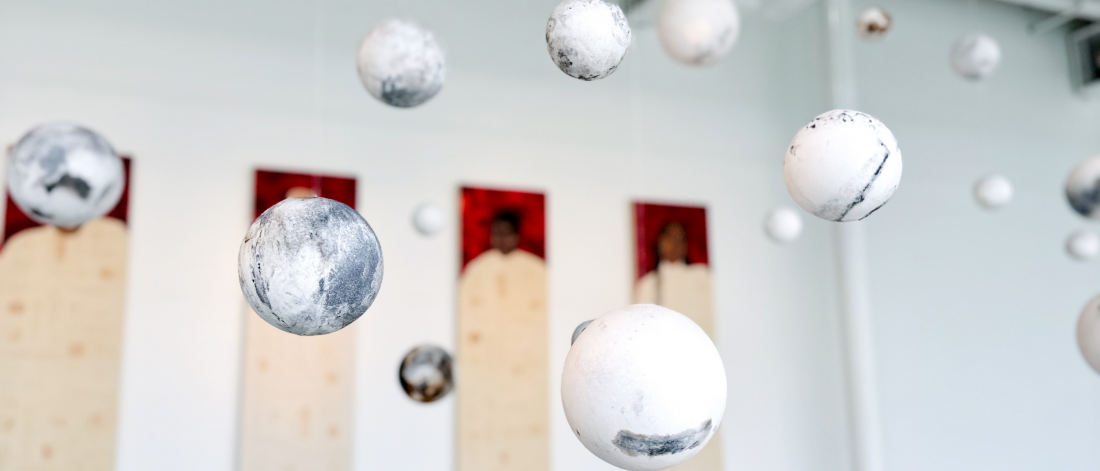 Installation of floating spheres in a gallery space with three red and beige portraits in the background.