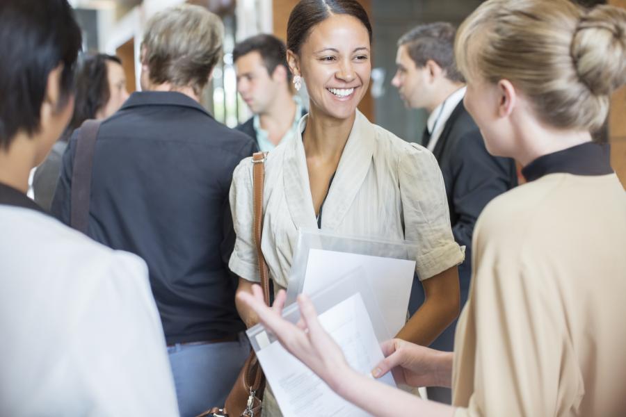Portrait of two smiling people talking, standing in crowded lobby.