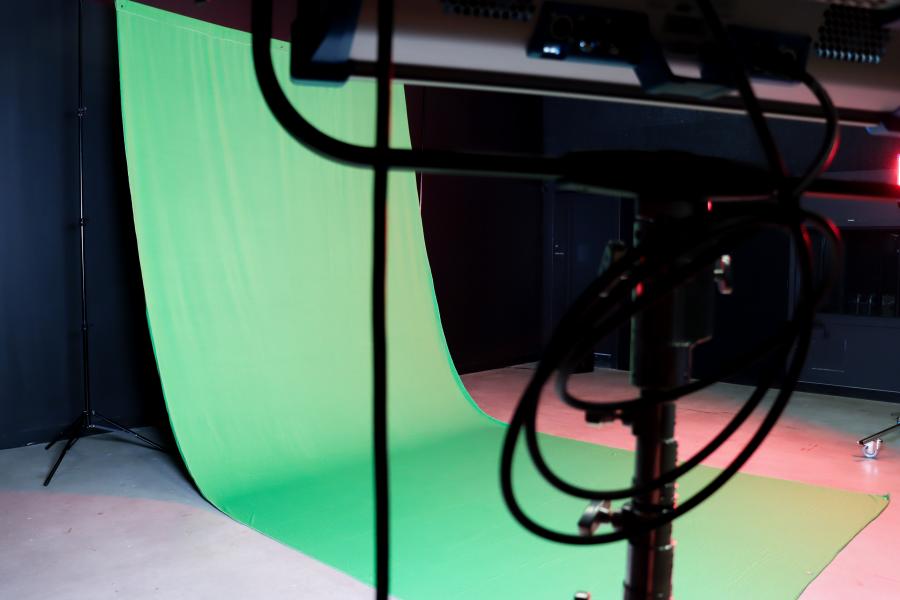 A professional video studio setup featuring a green screen backdrop, tripod-mounted camera, and studio lights in a dark-walled room.