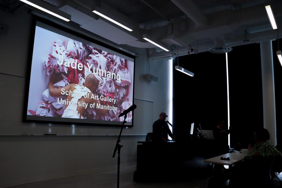An artist gives a talk in a darkened lecture room with a projected slide reading “Jade Yumang, School of Art Gallery, University of Manitoba.”