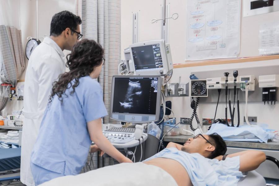 Two medical professionals look at a monitor while standing beside a patient in a bed.