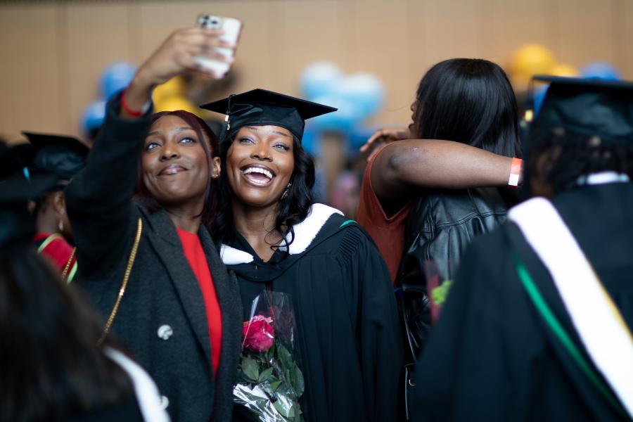 Two women smiling and posing for a selfie. Woman on the right is wearing graduation cap and gown, woman on the left is holding phone.